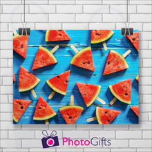 Load image into Gallery viewer, White painted brick wall with a landscape picture hanging from two clips. The picture is of some weatherbeaten wooden boards that have been painted a sky blue with triangles of water melon on lolly sticks all over the boards. As produced by Photogifts.co.uk
