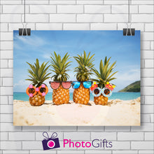 Load image into Gallery viewer, White painted brick wall with a landscape picture hanging on the wall from two clips. The picture is of four pineapples sitting on a sandy beach with blue sky and ocean in the background and the start of a small green hill in the back. All four pineapples are wearing different types of funny sunglasses. As produced by Photogifts.co.uk
