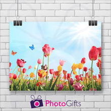 Load image into Gallery viewer, Landscape picture of a field of tulips in pinks, reds and yellows with butterflies flying around and a pale blue sky with the sun in the top right hand corner. The picture is hanging by two metal clips on a background of a white painted brick wall. As produced by Photogifts.co.uk
