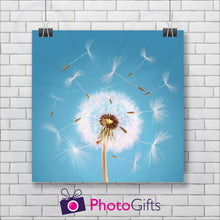 Load image into Gallery viewer, Square poster print with a close up shot of a dandelion seed head starting to come apart in the wind on a sky blue background. The picture is hung by two metal clips on a white painted brick wall. As produced by Photogifts.co.uk