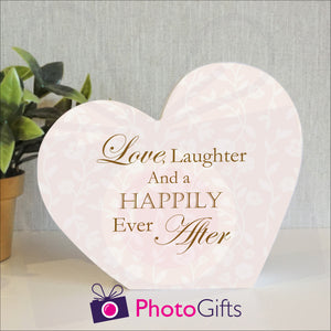 White wooden block in the shape of a heart with the slogan "Love, laughter and a happily ever after" printed on the block. The block and a potted plant is on a white shelf. Personalised block as supplied by Photogifts.co.uk