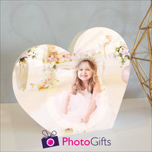 Load image into Gallery viewer, White wooden heart shaped block with a picture of a girl in a tiara and white dress sitting surrounded by flowers. White block and personalised photo as supplied by Photogifts.co.uk