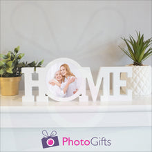 Load image into Gallery viewer, "Home" in a white painted wooden block with a picture of a mother and daughter having fun in stuck in the middle of the "O" of the home word. Shown on a white shelf with a couple of potted plants either side. Wooden word "Home" and personalised photo as supplied by Photogifts.co.uk