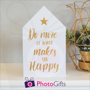 White wooden block in the shape of a house with the slogan "Do more of what makes you happy" printed on the front. The block is resting on a white shelf with a potted plant to one side. Personalised block as supplied by Photogifts.co.uk