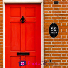 Load image into Gallery viewer, Red brick house with a close up of the red front door with black in door letterbox and knocker. To the right of the door as you face the door is a black oval panel with the number 82 and the word Smith printed on the panel along with some scroll work in the background. Panel as produced by Photogifts.co.uk