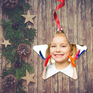 Dark wooden background with a Christmas Garland stretched out on the Left hand side from top to bottom. On the garland are some wooden stars and fir cones along with some dark red berries. To the right of the garland is a Christmas Decoration in the shape of a star with a red ribbon. The decoration is is a picture of a girl holding up her hands with each finger covered in a different colour paint as if she is about to do some hand painting.. Decoration as supplied by Photogifts.co.uk