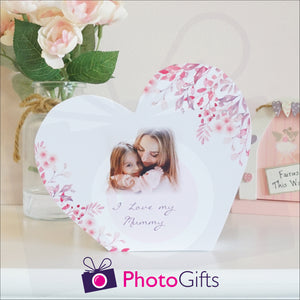 White wooden block in the shape of heart with the personalised photo of a mother and child with the words "I Love you mummy" and some flowers on a white shelf as supplied by Photogifts.co.uk