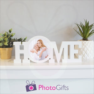 "Home" in a white painted wooden block with a picture of a mother and daughter having fun in stuck in the middle of the "O" of the home word. Shown on a white shelf with a couple of potted plants either side. Wooden word "Home" and personalised photo as supplied by Photogifts.co.uk