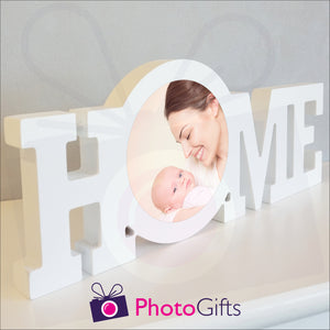 "Home" in a white painted wooden block with a picture of a mother and baby having fun stuck in the middle of the "O" of the home word. Shown on a white shelf as supplied by Photogifts.co.uk