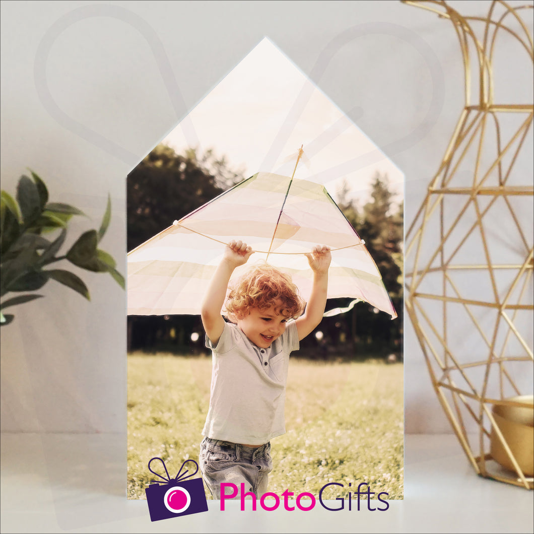 White wooden block in the outline of a house with a personalised photo of a boy and his kite attached to the block on a white shelf with a plant and a candle holder on either side. Block and personalised photo as supplied by Photogifts.co.uk