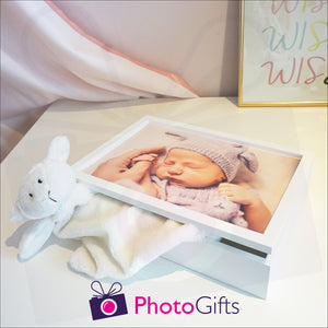 White wooden keepsake box that has been personalised with a baby's photo holding on to a parents hand. Box is slightly open with a soft toy partially in the box. The box is sitting on a white shelf with a curtain and picture in the background. Personalised box as supplied by Photogifts.co.uk