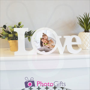 "Love" in a white painted wooden block with a picture of a mother and daughter having fun in stuck in the middle of the "O" of the love word. Shown on a white shelf with a couple of potted plants either side. Wooden word "Love" and personalised photo as supplied by Photogifts.co.uk