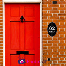 Load image into Gallery viewer, Red brick house with a close up of the red front door with black in door letterbox and knocker. To the right of the door as you face the door is a black oval panel with the number 82 and the word Smith printed on the panel along with some scroll work in the background. Panel as produced by Photogifts.co.uk
