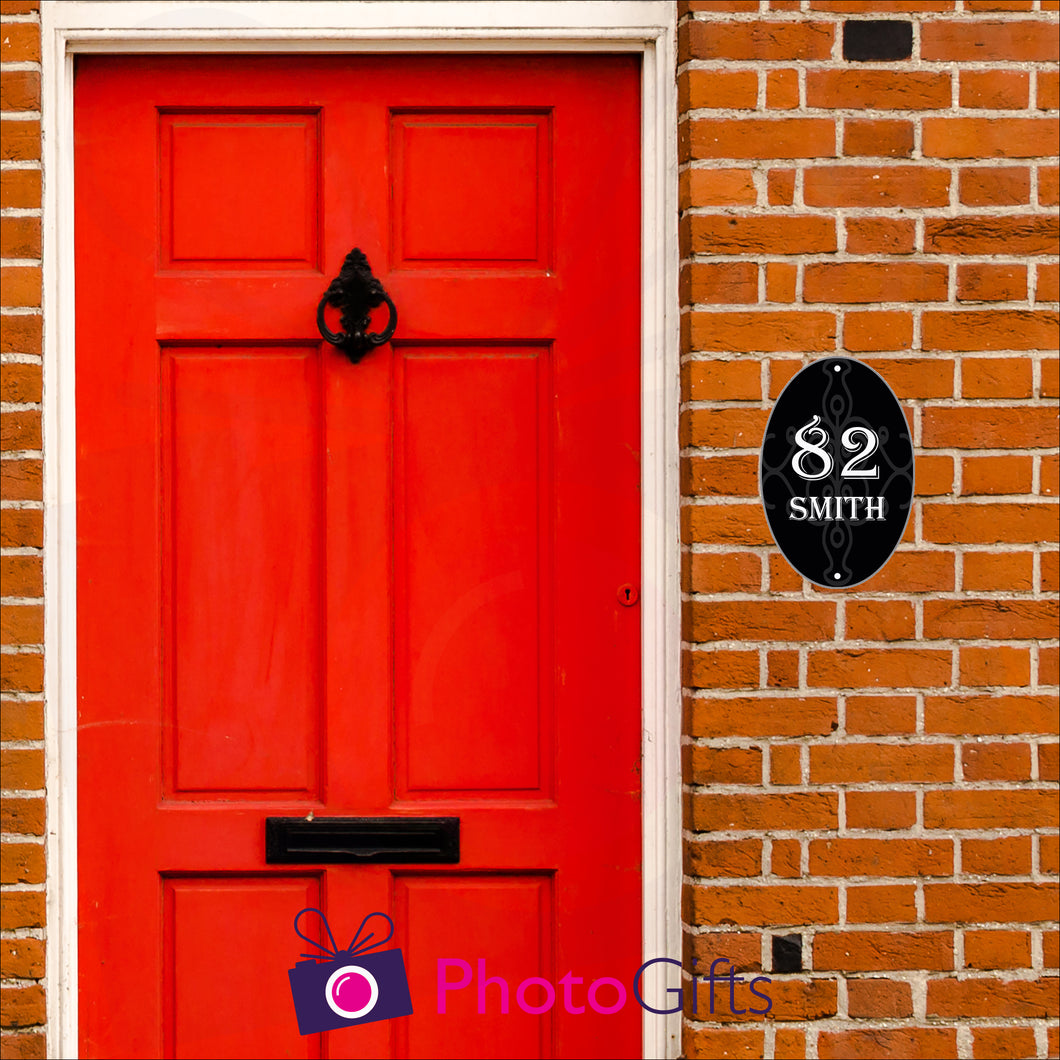 Red brick house with a close up of the red front door with black in door letterbox and knocker. To the right of the door as you face the door is a black oval panel with the number 82 and the word Smith printed on the panel along with some scroll work in the background. Panel as produced by Photogifts.co.uk