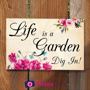 Rectangular panel in landscape orientation on a wooden fence. On the panel is the slogan "Life is a garden Dig In!" together with pictures of some vibrant flowers and some bright blue butterflies. As produced by Photogifts.co.uk
